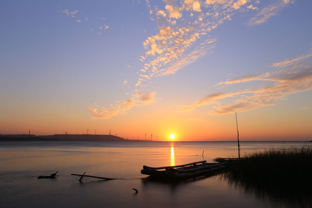 Beautiful sunrise over a lake featuring a wooden boat and distant windmills under a vibrant sky.